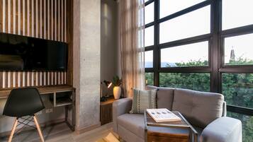 Una sala de estar moderna con un sofá gris, un escritorio de madera, una silla negra y un ventanal con vistas a follaje y una estructura distante.