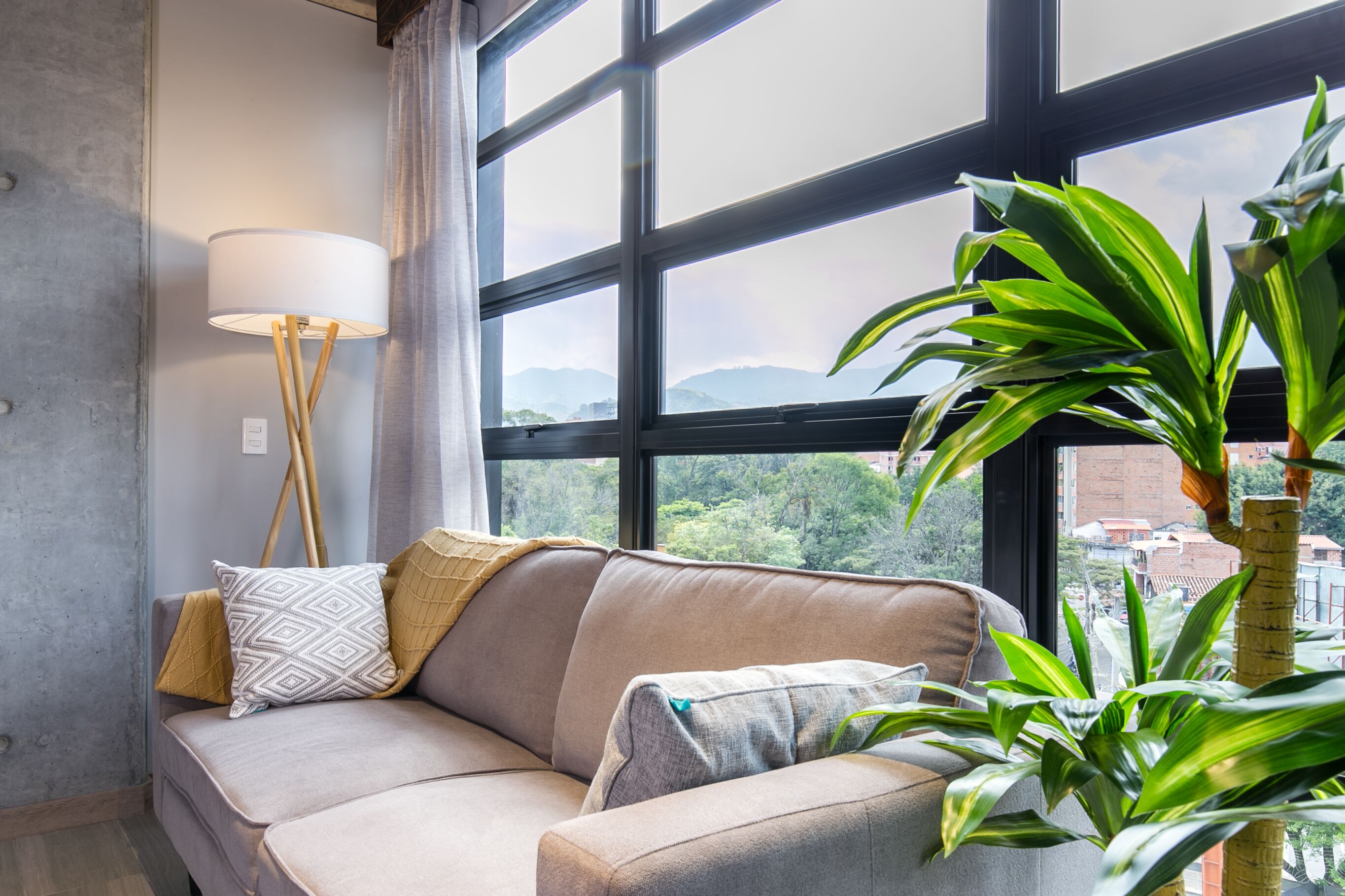 A living room with a sofa, a floor lamp, and a large window with a view of greenery and buildings.