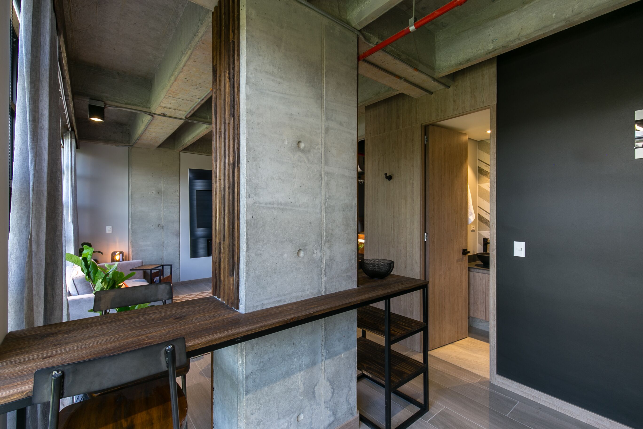 Een modern interieur met een betonnen accentmuur, een houten bank en een keukenruimte zichtbaar door een open deur.