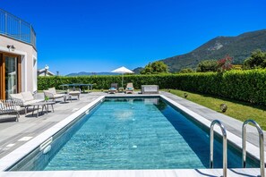 Uma piscina retangular com superfície de água azul cristalina, rodeada por um deck ladrilhado. Há espreguiçadeiras e uma mesa pequena com um abajur no deck. Um guarda-sol branco é visível ao fundo.