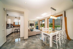 Una cocina moderna con encimera de mármol y gabinetes blancos, una zona de comedor con mesa de vidrio y sillas blancas, y un salón con sofá y televisor.