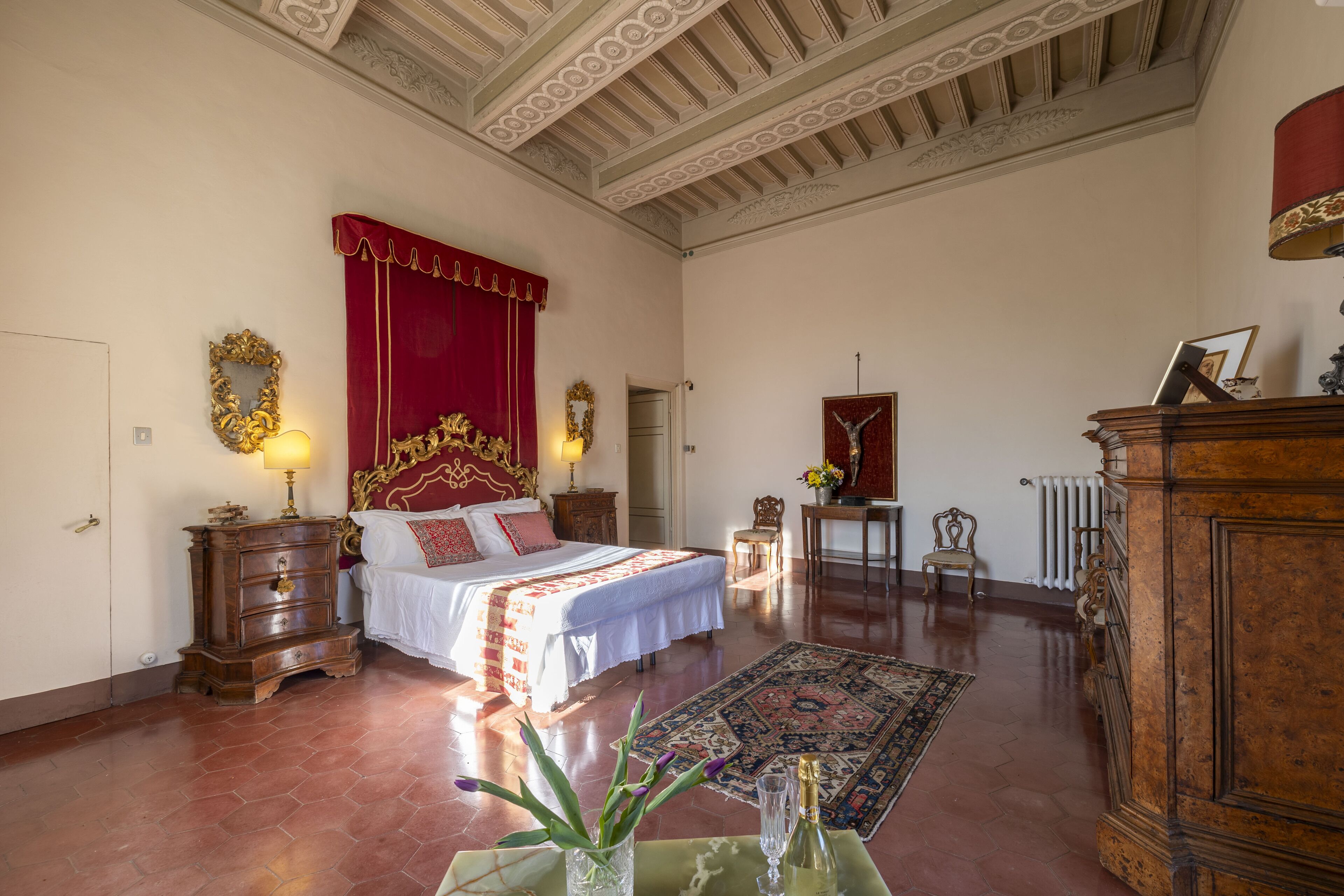 Un dormitorio espacioso con una cama grande, un dosel rojo, un aparador de madera y una lámpara de araña clásica.