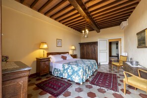A bedroom with a wooden ceiling, a bed with a floral bedspread, two bedside lamps, a wooden nightstand, a red and black rug, and a wooden chair. - CASTELLO DI SOVICILLE- RESIDENZA D'EPOCA (Sovicille)