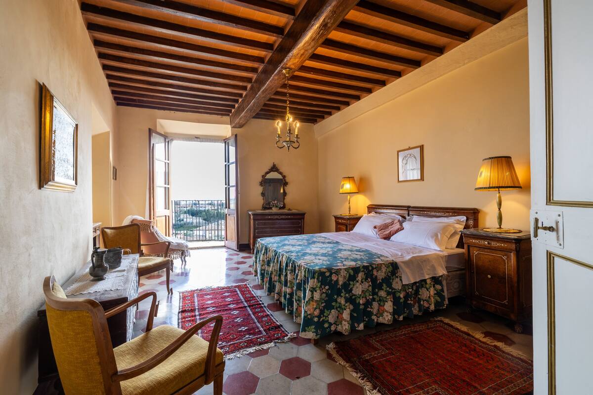 A bedroom with a wooden ceiling, a bed with a floral bedspread, a nightstand with a lamp, a chair, a table with a pitcher, and a view through an open door.