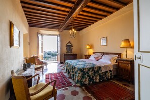 A bedroom with a wooden ceiling, a bed with a floral bedspread, a nightstand with a lamp, a chair, a table with a pitcher, and a view through an open door. - CASTELLO DI SOVICILLE- RESIDENZA D'EPOCA (Sovicille)