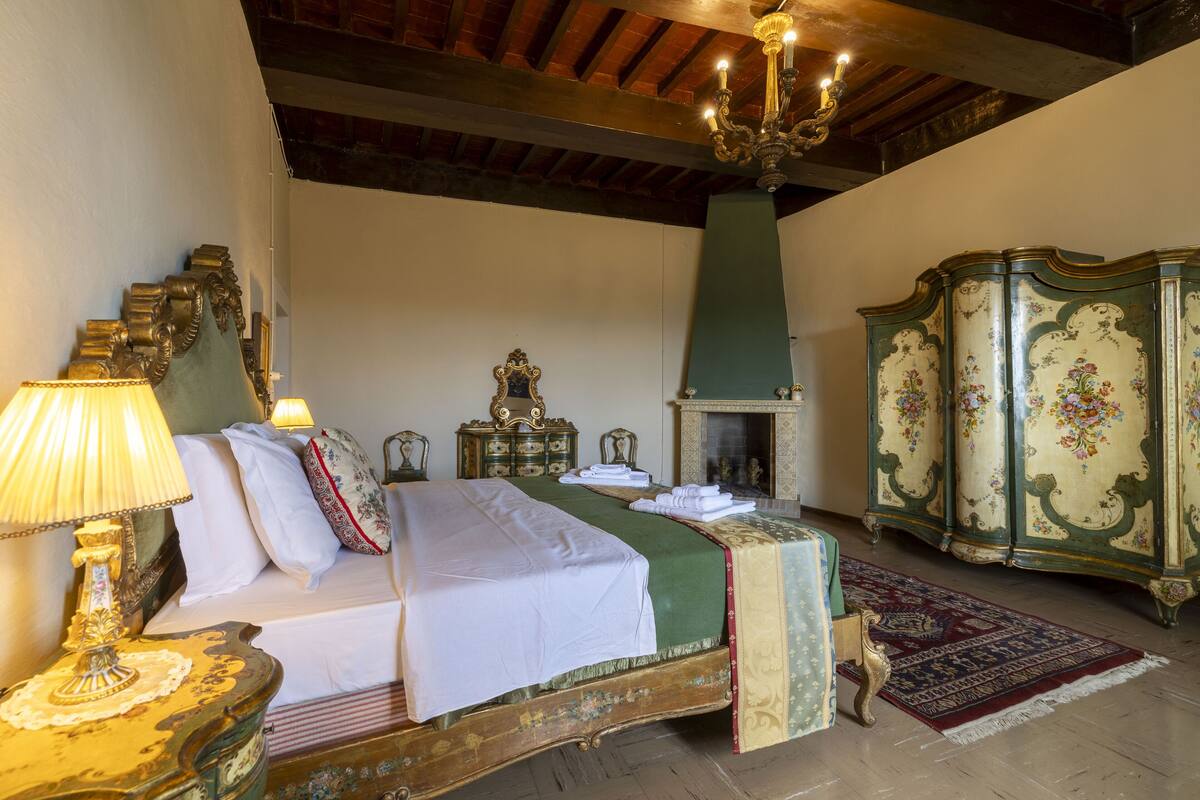 A traditional room with a bed, a chandelier, a fireplace, and a green cabinet.