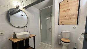 Kylpyhuone, jossa on puinen allaskaappi, pyöreä peili, wc ja lasiseinällä varustettu suihku.