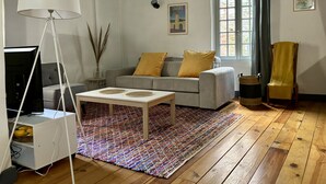 Olohuoneessa on sohva, kaksi keltaista koristetyynyä, valkoinen lamppu, valkoisella jalustalla oleva televisio, värikäs matto, ikkuna, jossa on verhot, ja seinällä oleva juliste.