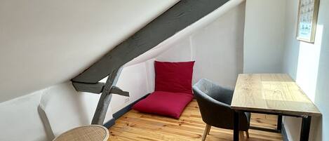 Ein kleiner, gemütlicher Raum mit schrägverdeckter Decke, Holzboden, einem Tischchen, einem Stuhl, einem roten Kissen und einem Korbstuhl.