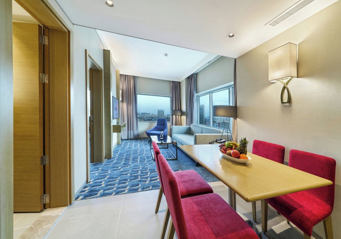 Habitación de hotel con comedor, sofá, mesa con fruta y vista a la ciudad.
