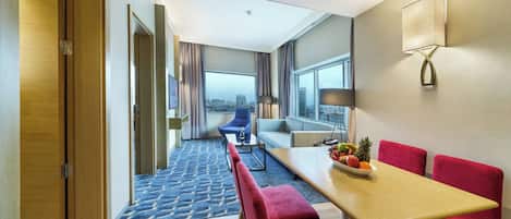 Habitación de hotel con comedor, sofá, mesa con fruta y vista a la ciudad.