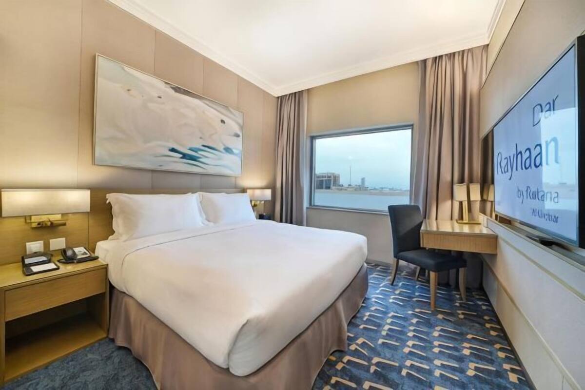 A hotel room with a large bed, a desk, a chair, a TV, and a view of the city.