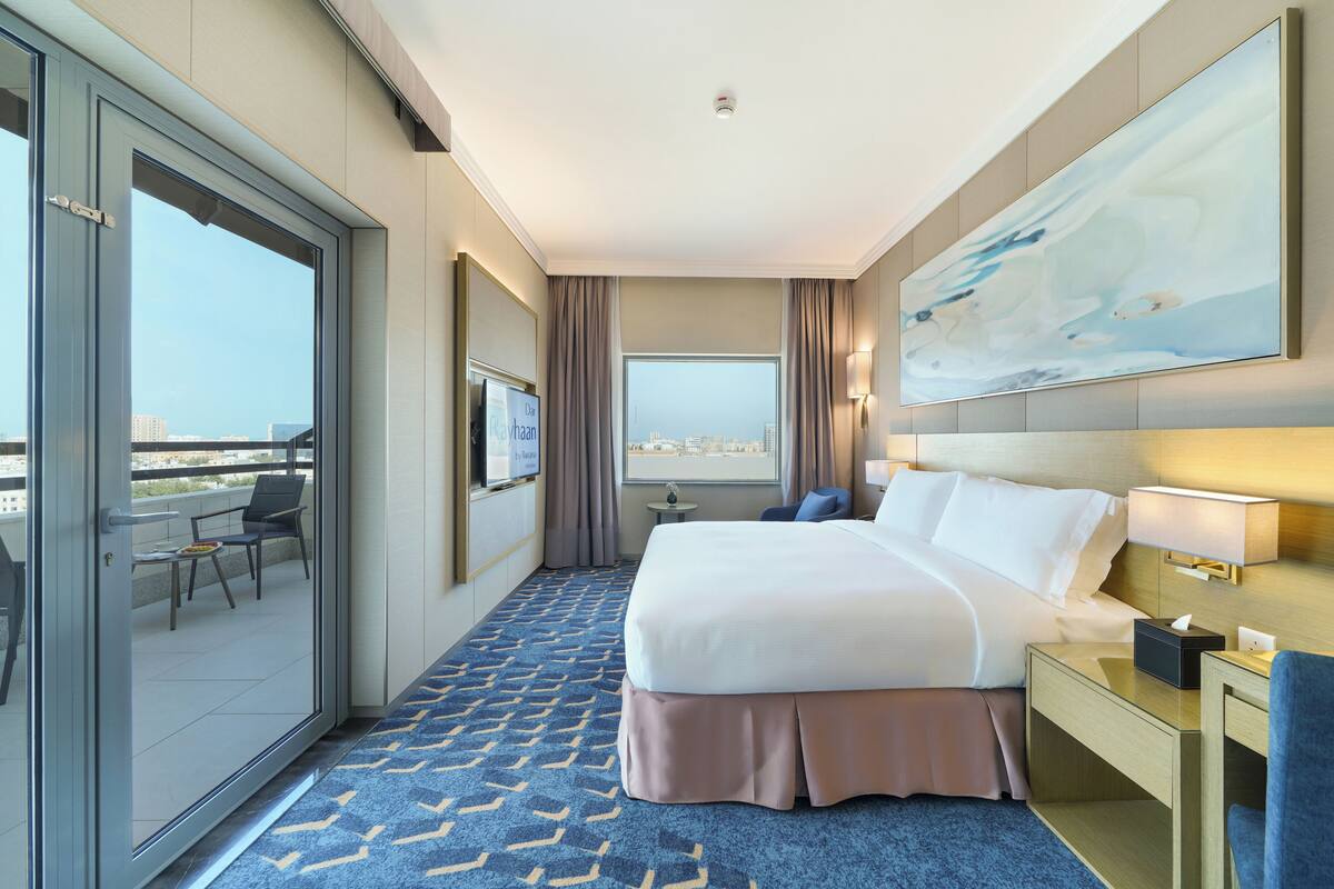 A hotel room with a bed, a desk, a chair, a TV, and a balcony with a view of the city.