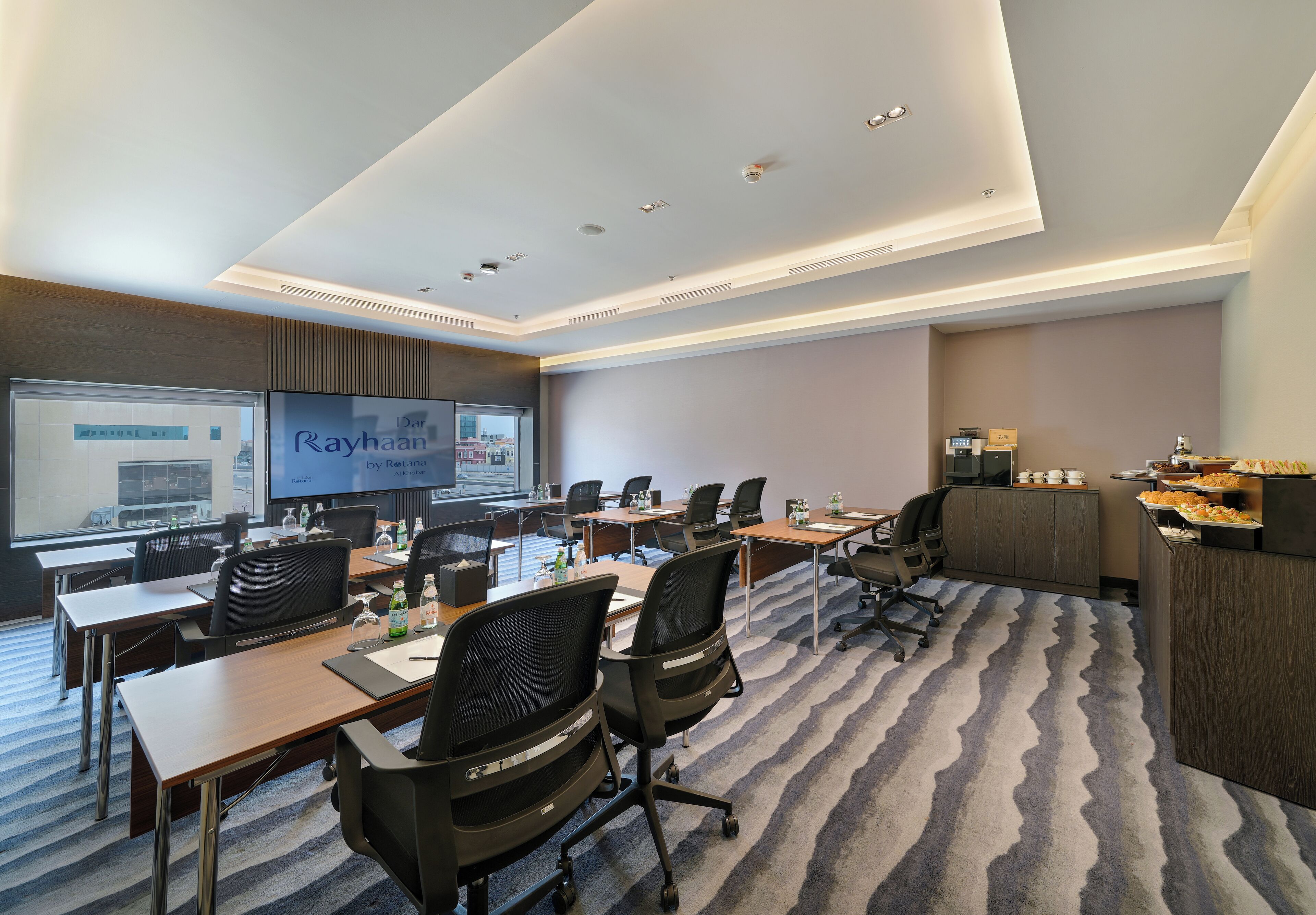 A conference room with multiple tables, chairs, a large screen, and a buffet area.