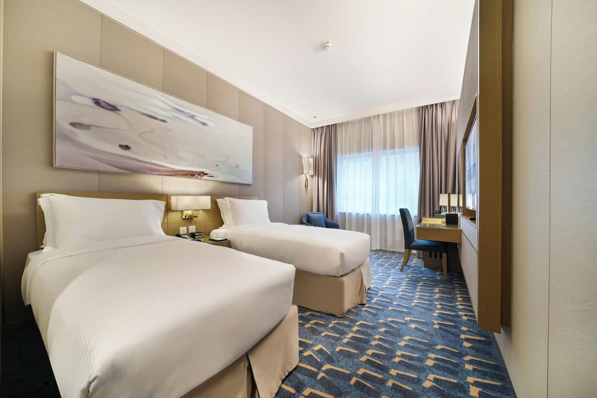A hotel room with two beds, a desk, a chair, a large abstract painting, and a patterned carpet.