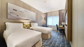 A hotel room with two beds, a desk, a chair, a large abstract painting, and a window with curtains.