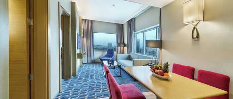 A hotel room with a dining area, a sofa, a blue chair, and a view of the city.