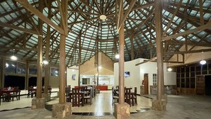 Un vaste espace intérieur doté d’un haut plafond soutenu par des poutres en bois et surmonté d’une toiture en verre. Des tables et des chaises sont disposées pour les repas.
