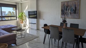 En moderne stue med en sofa, et spisebord med stole, et tv ophængt på væggen og et stort vindue med udsigt til bygninger og træer.