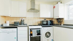 A compact kitchen with white cabinets, a gas stove, a microwave, a toaster, a kettle, and a washing machine. - Canterbury Charm, Spacious 4 Bed House (Canterbury)