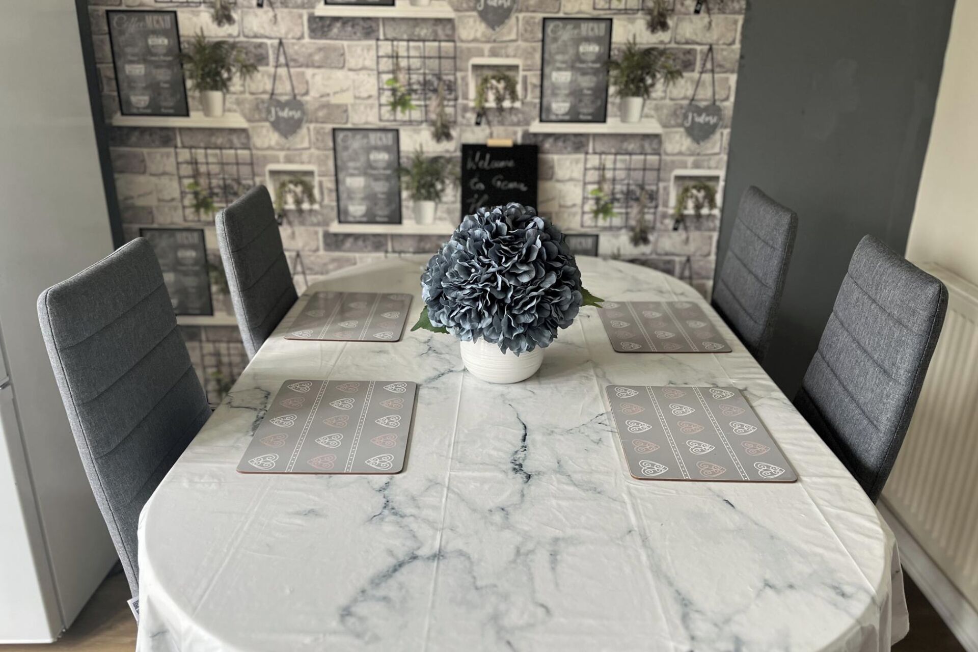 A dining table with a marble top, surrounded by grey upholstered chairs. A potted plant with dark blue flowers is centered on the table.