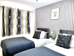 Two beds with dark blue bedding and a white headboard, a nightstand with a lamp and a potted plant, and a window with curtains.
