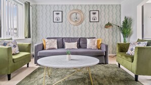 Uma sala de estar com um sofá cinza, poltronas verdes, uma mesa de centro redonda e papel de parede estampado.