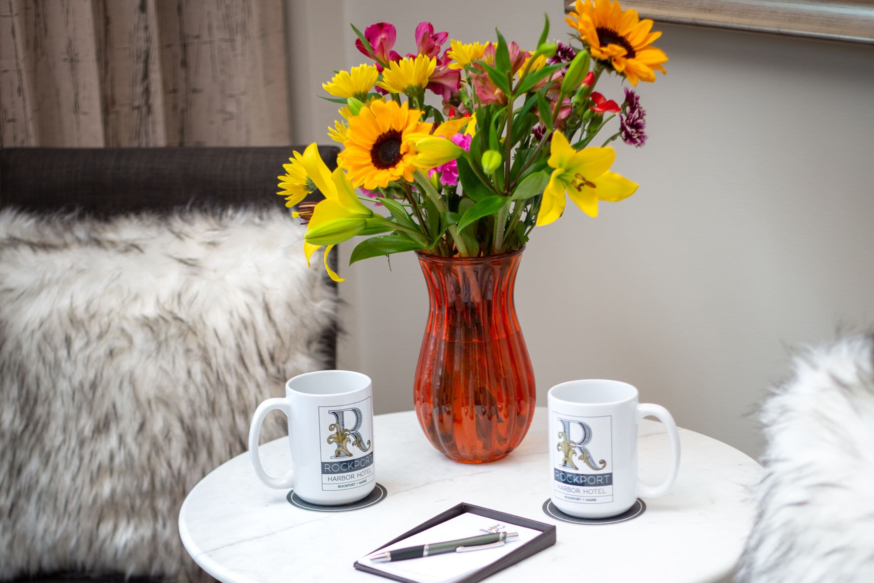 A white table with a vase of colorful flowers, two mugs with a logo, and a pen on a white surface.