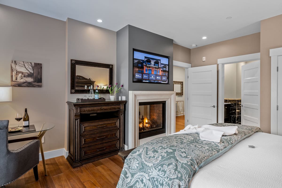 A bedroom with a fireplace, a TV mounted above it, a bed with a patterned bedspread, a desk with a chair, and a mirror.