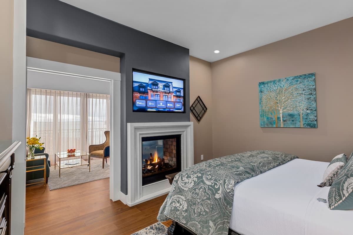 A bedroom with a fireplace, a bed with patterned bedding, a television mounted above the fireplace, and a painting on the wall.