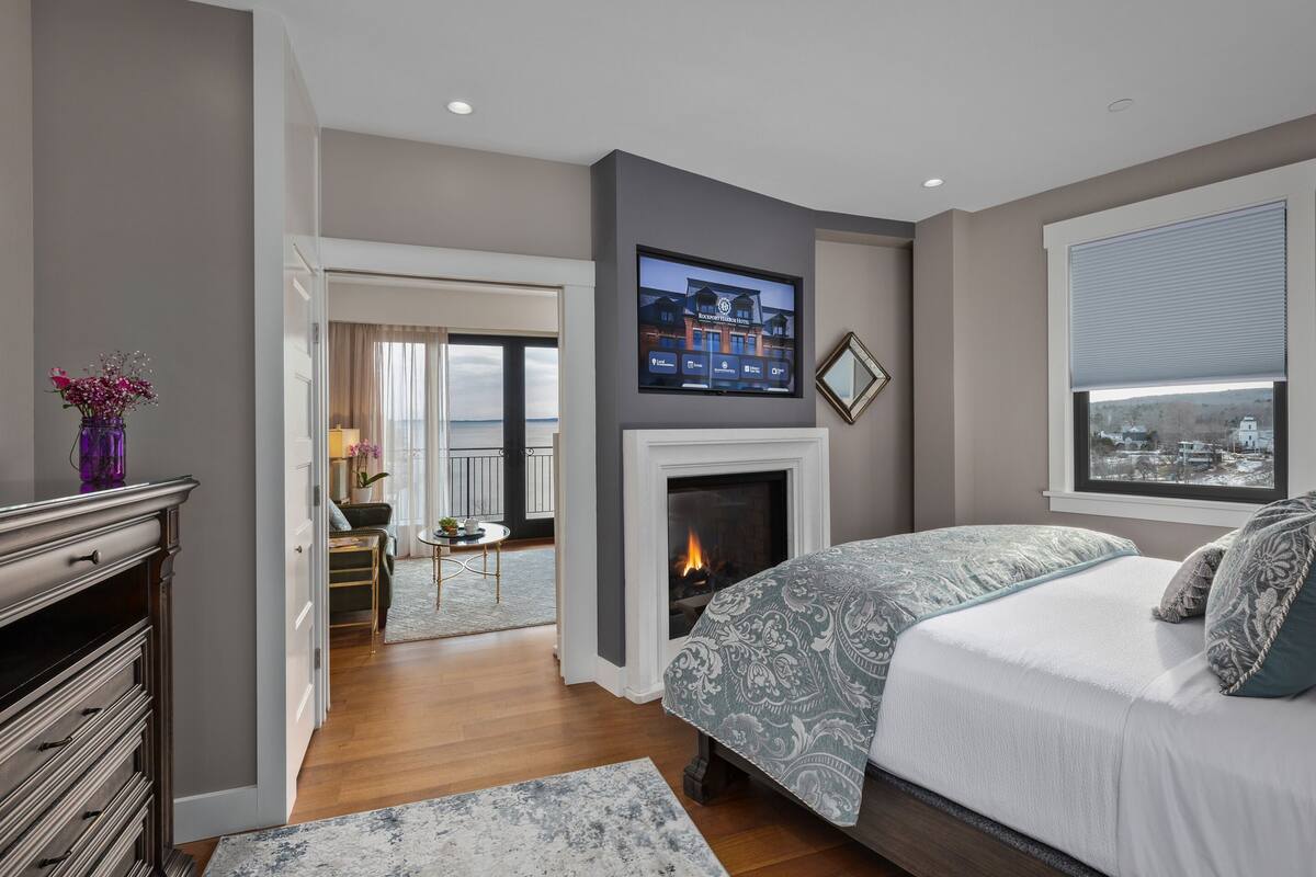 A modern hotel room with a fireplace, a bed with patterned bedding, a television mounted above the fireplace, and a view of the city through a window.