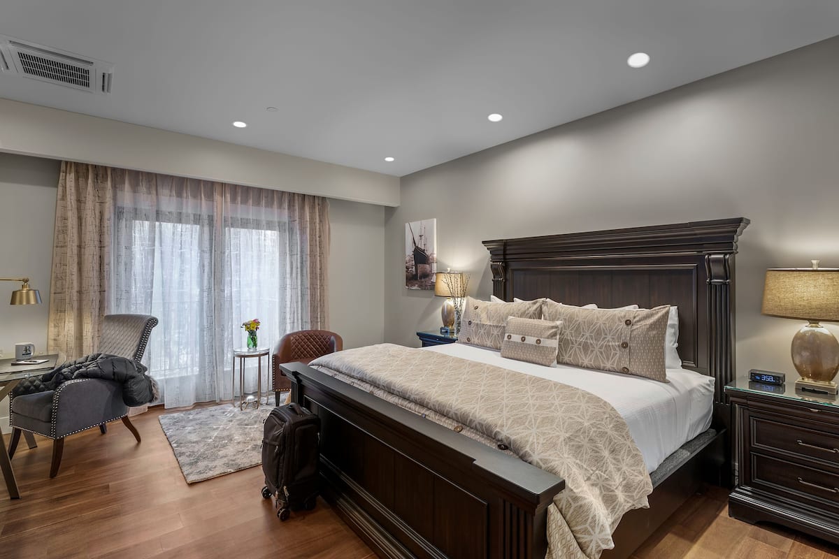 A bedroom with a large bed, a desk, a chair, and a small table with a lamp.