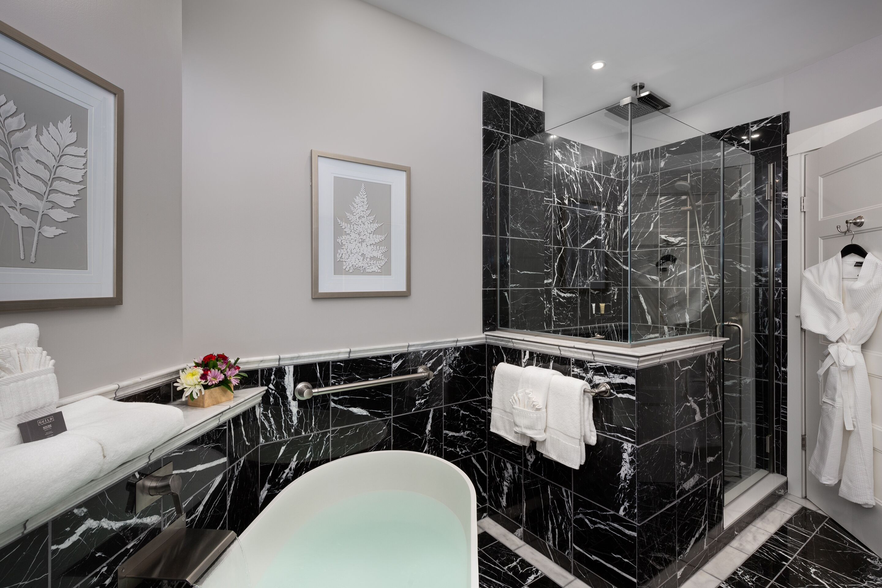 A modern bathroom with a marble vanity, a freestanding bathtub, a glass-enclosed shower, and a white robe hanging on the wall.