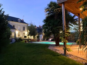 Un jardin bien entretenu avec une piscine, une grande maison dotée de nombreuses fenêtres et une pergola en bois.