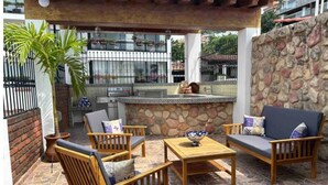 Un patio al aire libre con piso de piedra, mobiliario de madera, pared de piedra y una zona de bar con parrilla.