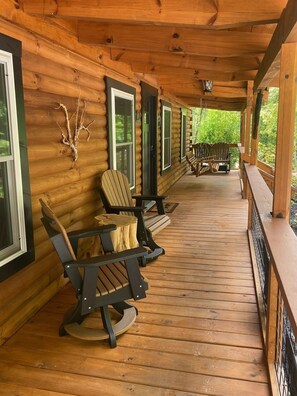 Puinen mökki, jossa on kuisti, Adirondack-tyyliset puutarhatuolit, puinen terassi ja näkymä metsään.