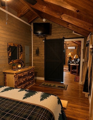 A cozy log cabin interior with a bed, wooden dresser, and a television mounted on the wall.