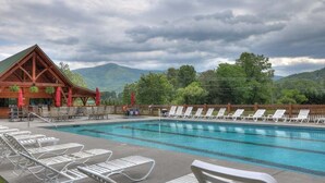 A swimming pool with lounge chairs, a wooden building with a green roof, and mountains in the background.