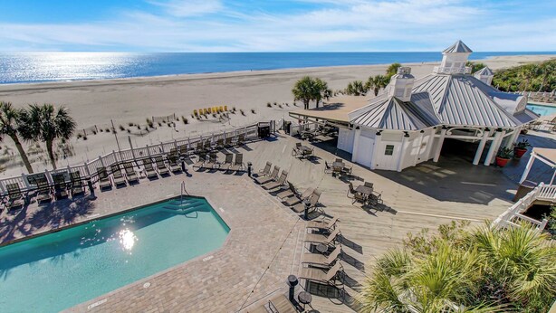 A pool area with lounge chairs and a small building with a unique roof design. - 131 Grand Pavilion by Avantstay Oceanfront On The Boardwalk! (Isle of Palms)