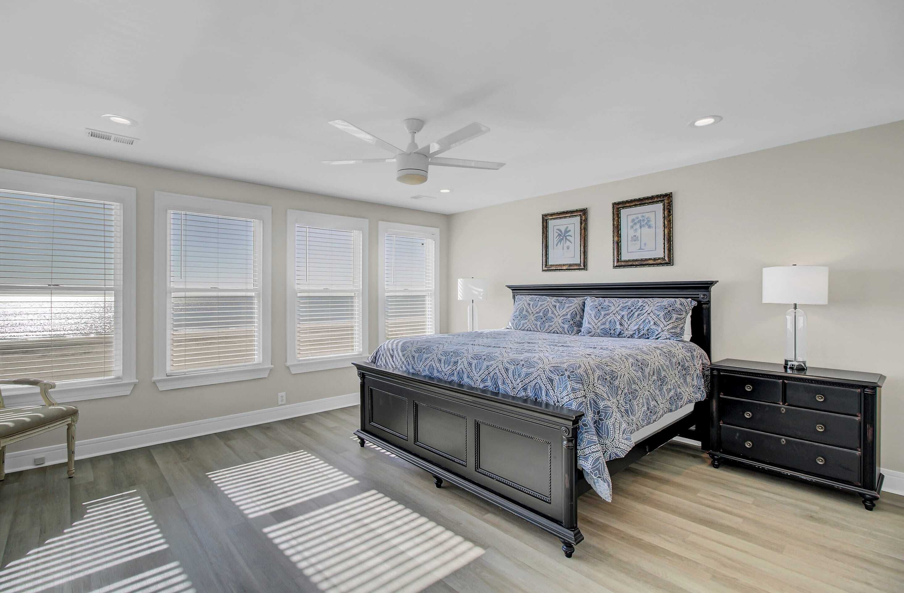 Une chambre à coucher comprenant un lit, des tables de chevet, une commode, un ventilateur de plafond et de grandes fenêtres équipées de stores.