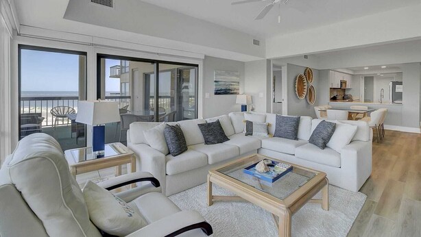 Een ruime woonkamer met een grote witte bank, een glazen salontafel en uitzicht op de oceaan door grote schuifdeuren.