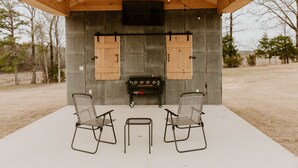 An outdoor patio with a grill, two chairs, and a television screen on a wooden ceiling.