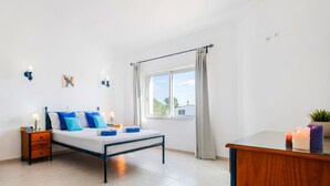 A bedroom with a bed, nightstand, bedside table, lamp, curtains, and a window.