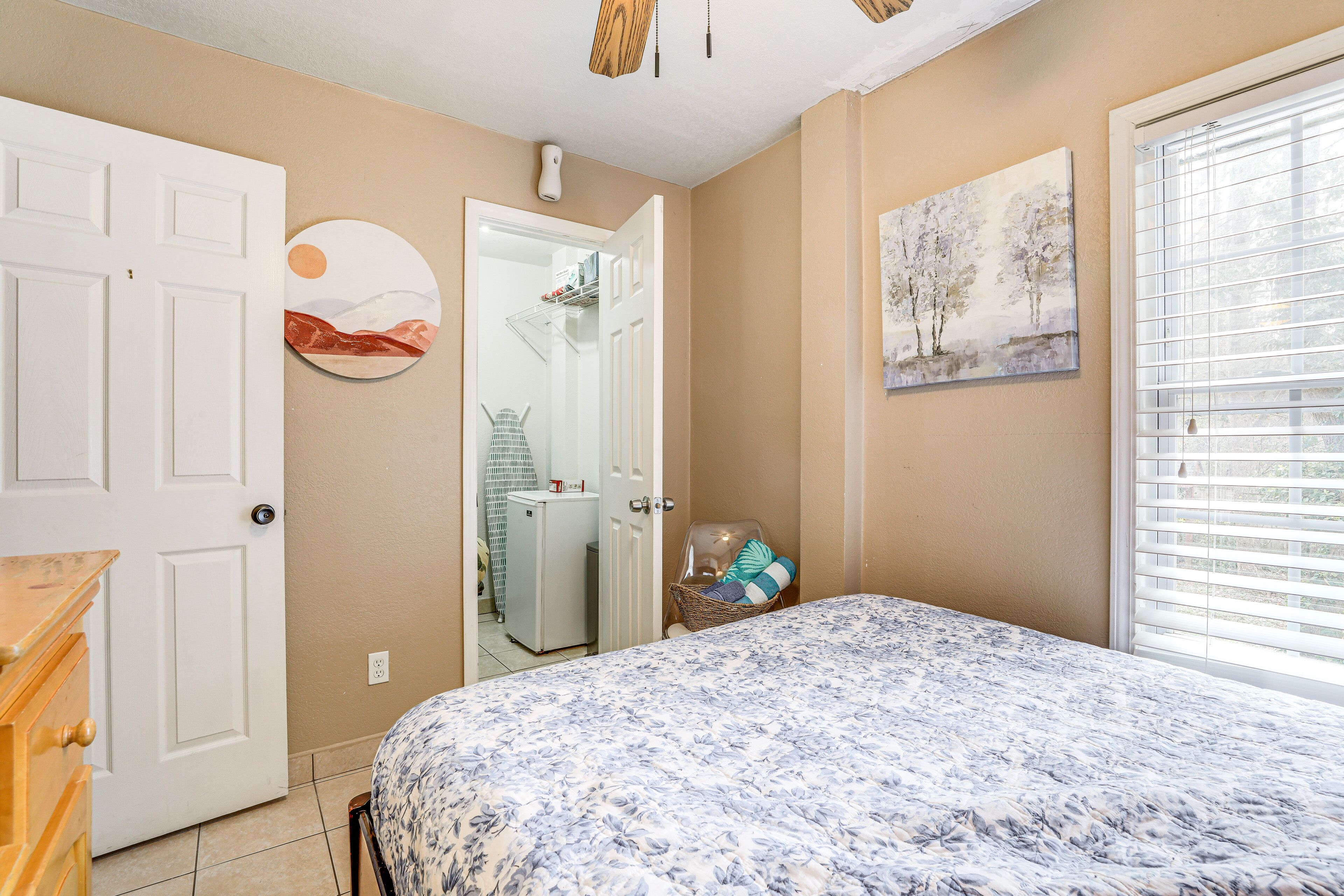 A bedroom with a bed, a door, a wall-mounted painting, and a window with blinds.