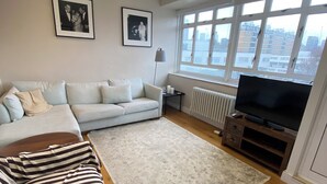 A living room with a sofa, a striped blanket, a TV, and a window with a view of buildings. - Delightful & Well-located 2BD Flat Central Pimlico (London)
