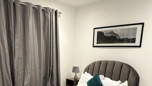 Ein ordentlich bezogenes Bett mit einer grünen Tagesdecke, einem grauen Kopfteil und weißen Kissen. Ein kleiner Nachttisch mit Lampe und ein gerahmtes Bild an der Wand sind ebenfalls zu sehen.