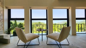 Ein Zimmer mit zwei Stühlen und einem kleinen Tisch, große Fenster mit Blick auf Grünflächen und Hügel.
