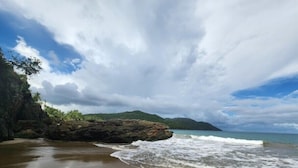 Ein Strand mit einem Felsen, Wellen und einem entfernten Berg unter bewölktem Himmel.