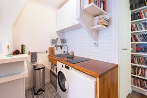 A compact kitchen with a washing machine, a sink, and a bookshelf filled with books. - Bright Stylish 1 BDR Flat Folies Bergere (Paris)