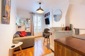 A compact living space with a sofa, a small table, a chair, a TV, a mirror, and a kitchen area with a countertop and cabinets.